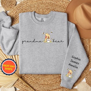 May include: A light gray sweatshirt with the words "grandma bear" in cursive, and a cartoon bear with bees. The sleeve has the names Sophia, James, and Amelia. A round sticker with "NEXT DAY SHIPS" is visible.