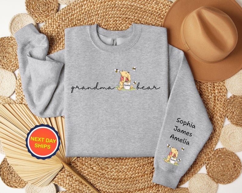 May include: A light gray sweatshirt with the words "grandma bear" in cursive, and a cartoon bear with bees. The sleeve has the names Sophia, James, and Amelia. A round sticker with "NEXT DAY SHIPS" is visible.