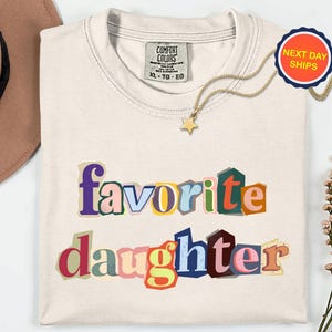 May include: Cream-coloured t-shirt with the words "favorite daughter" in colourful, cut-out style letters. A gold necklace with a star pendant and a "Next Day Ships" sticker are also visible.
