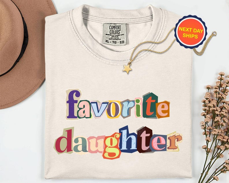 May include: Cream-coloured t-shirt with the words "favorite daughter" in colourful, cut-out style letters. A gold necklace with a star pendant and a "Next Day Ships" sticker are also visible.