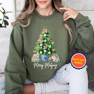 May include: Olive green sweatshirt featuring a Christmas tree design with pink bows, ornaments, and wrapped gifts. The words "Merry Mahjong" are written in white script below the tree. A round sticker with "Next Day Ships" is visible.