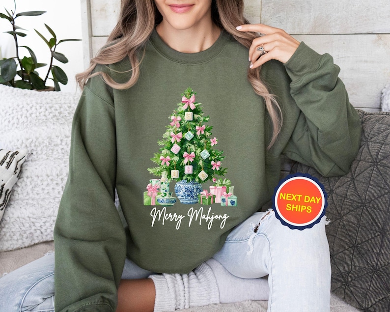 May include: Olive green sweatshirt featuring a Christmas tree design with pink bows, ornaments, and wrapped gifts. The words "Merry Mahjong" are written in white script below the tree. A round sticker with "Next Day Ships" is visible.