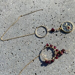 May include: Two gold hoop earrings with dangling charms. The first earring has a round charm with a sun and moon design. The second earring has a circle of small red beads.