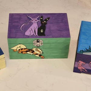May include: Three rectangular wooden boxes with metal latches, each featuring hand-painted characters. The left box is blue and yellow with Meowth and Pikachu. The centre box is purple and green with Espeon, Umbreon, and Arcanine. The right box is blue and purple with Dragonite and Mew.