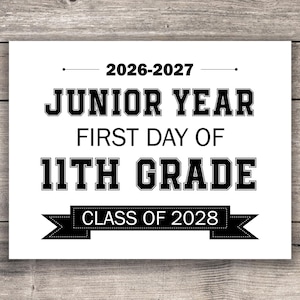 May include: A white sign with black text on a wooden background. The sign reads "2026-2027 JUNIOR YEAR FIRST DAY OF 11TH GRADE CLASS OF 2028". The text is in a bold, collegiate font.