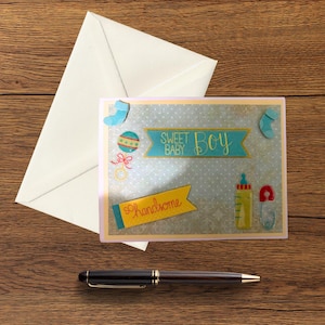 May include: A baby shower greeting card with the words "Sweet Baby Boy" and "so handsome" on it. The card has illustrations of baby items like a bottle, rattle, and socks. A white envelope and a pen are also on the wooden surface.