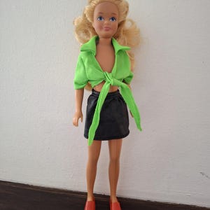 以下が含まれることがあります： ブロンドの髪と青い目のヴィンテージドール。緑色の結び目のトップスと黒いスカートを着用。赤い靴を履いて白い壁の前に立っています。