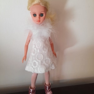 Könnte beinhalten: Eine weiße Puppe mit blonden Haaren, die ein weißes Kleid mit einem weißen Pelzkragen und rosa Glitzer-Stiefeln trägt.