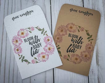 May include: Two brown paper seed packets with a floral design and the text "Bloom with Baby Lia" printed on them. The packets are labeled "Wildflower Seeds".