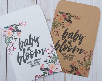 May include: Two white and brown paper favor bags with floral designs and the text "baby in bloom" and "DANIELLE & DARYL'S BABY SHOWER MAY 6, 2018".