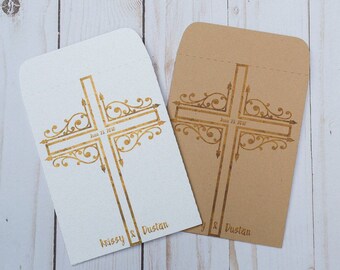 May include: Two white and brown paper envelopes with a gold cross design. The cross has a decorative scroll design and the text "June 23, 2018" and "Krissy & Dustan".