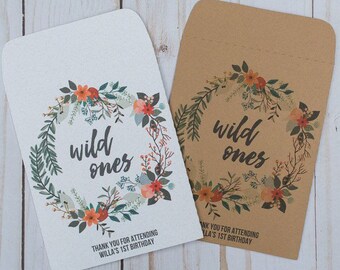 May include: Two brown paper favor bags with a floral wreath design and the text "wild ones". The bags have a thank you message for attending a first birthday party.