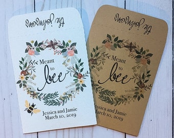May include: Two white and brown paper favor bags with a floral wreath design. The bags have the text "Meant to bee" and the names "Jessica and Jamie" with the date "March 10, 2019".
