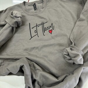 May include: A gray sweatshirt with the words "Let Them" in black script, a small red heart accents the text. The long sleeves are rolled up. The sweatshirt is made of a soft material.