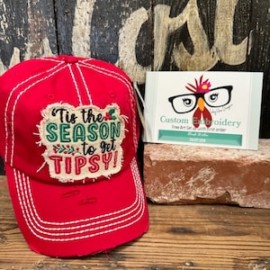 May include: Red baseball cap with a distressed white stitched brim and a green and red embroidered patch that says "Tis the Season to get Tipsy!"
