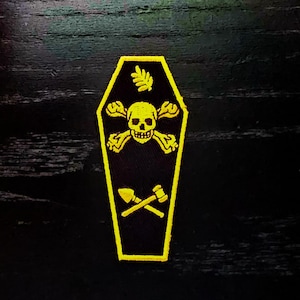 May include: Yellow embroidered patch shaped like a coffin with a skull and crossbones, a leaf, and two crossed tools inside. The patch is on a black background.