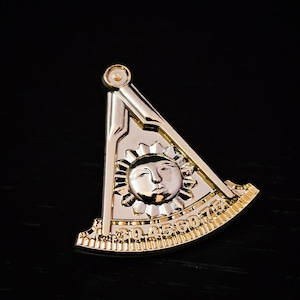 May include: A gold and silver-toned Masonic emblem featuring a sun with a face at the center, set within a compass and square design. The emblem has detailed engravings and is set against a dark background.