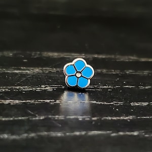 Puede incluir: Un pequeño pin de flor azul, no me olvides, con acabado plateado y un solo strass blanco en el centro.