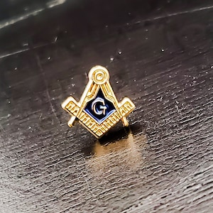 May include: A gold-colored Masonic pin featuring the Square and Compasses symbol with a blue center and the letter "G". The pin has a detailed design and is set against a dark background.