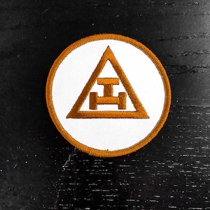 May include: A white embroidered patch with a brown outline. The patch features a triangle with a horizontal bar and two circles at the base.