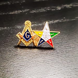 May include: A gold-tone metal lapel pin featuring two conjoined emblems: a Masonic square and compass with a blue center containing the letter G, and a five-pointed Order of the Eastern Star emblem with white, yellow, green, blue, and red enamel.