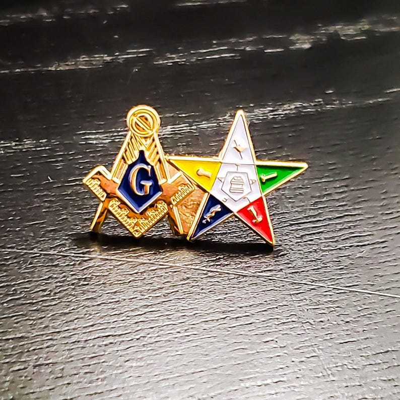 May include: A gold-tone metal lapel pin featuring two conjoined emblems: a Masonic square and compass with a blue center containing the letter G, and a five-pointed Order of the Eastern Star emblem with white, yellow, green, blue, and red enamel.
