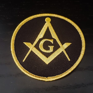 May include: A black and gold embroidered patch featuring a square and compass with a 'G' in the center. The patch is circular and has a gold border.