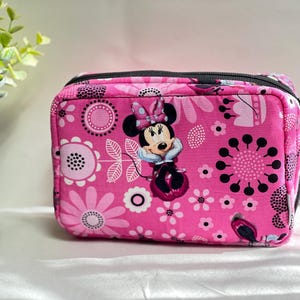 Op de afbeelding: Roze rechthoekig etui met ritssluiting met een bloemenpatroon en een afbeelding van Minnie Mouse. De etui heeft een zwarte ritssluiting en is versierd met witte en zwarte bloemen. De achtergrond is lichtroze.