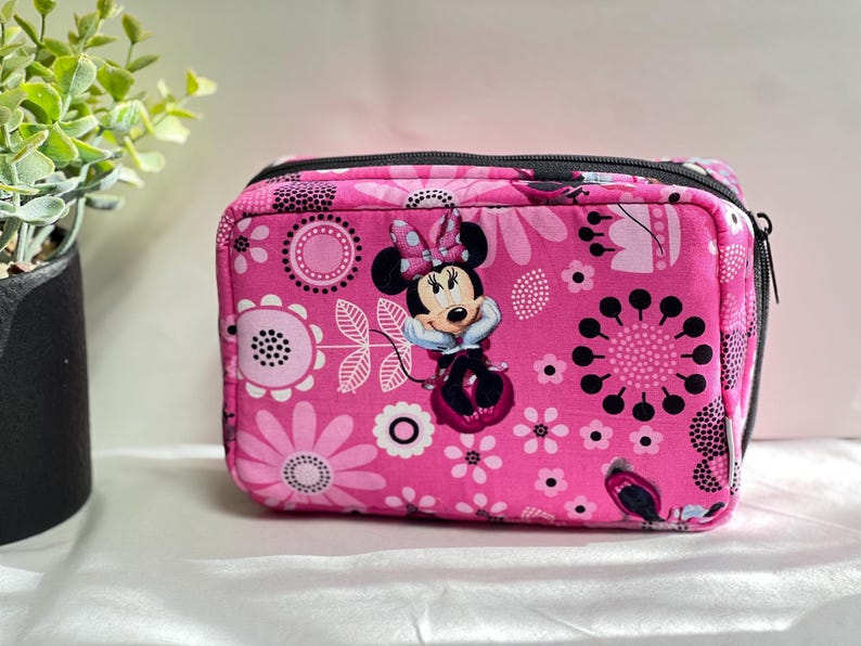 Op de afbeelding: Roze rechthoekig etui met ritssluiting met een bloemenpatroon en een afbeelding van Minnie Mouse. De etui heeft een zwarte ritssluiting en is versierd met witte en zwarte bloemen. De achtergrond is lichtroze.