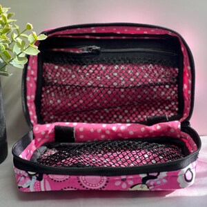 Op de afbeelding: Open roze reisetui met zwarte rand, met een bloemen- en stippenpatroon. De binnenkant heeft gaasvakken en een vak met ritssluiting. De etui is open en onthult de inhoud.