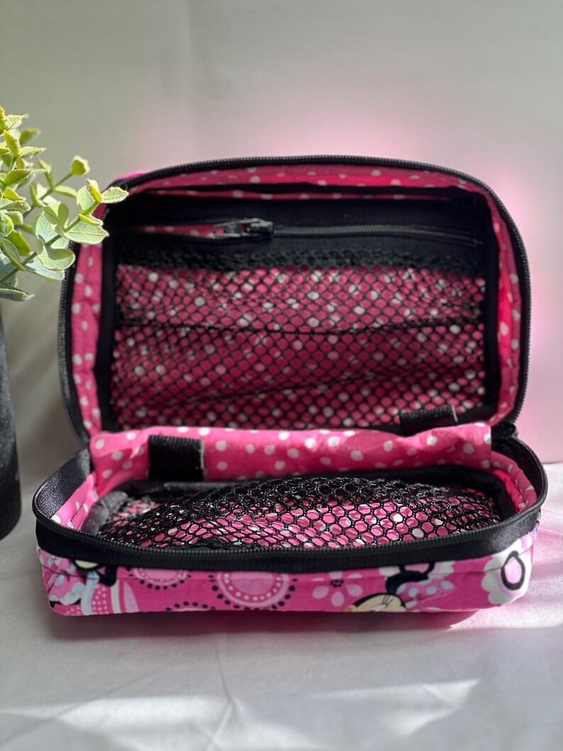 Op de afbeelding: Open roze reisetui met zwarte rand, met een bloemen- en stippenpatroon. De binnenkant heeft gaasvakken en een vak met ritssluiting. De etui is open en onthult de inhoud.