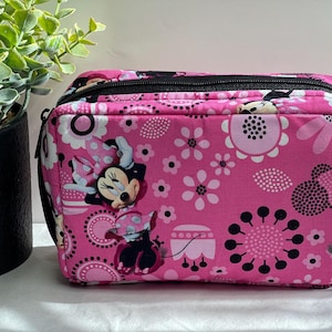 Op de afbeelding: Een rechthoekige roze reistas met een zwarte rits en handvat. De tas is versierd met een bloemenpatroon en afbeeldingen van Minnie Mouse. De tas staat op een wit oppervlak.