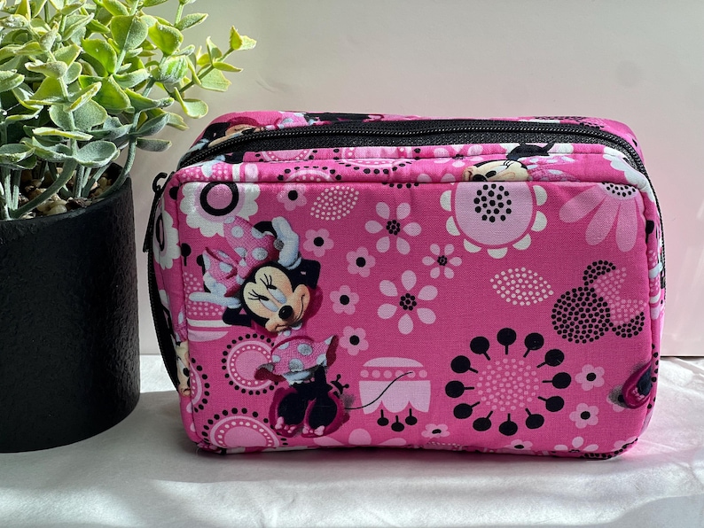 Op de afbeelding: Een rechthoekige roze reistas met een zwarte rits en handvat. De tas is versierd met een bloemenpatroon en afbeeldingen van Minnie Mouse. De tas staat op een wit oppervlak.