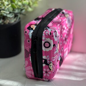 Op de afbeelding: Een rechthoekige roze make-up tas met een zwarte ritssluiting. De tas is versierd met een bloemenpatroon en stripfiguren. De tas is gemaakt van stof.