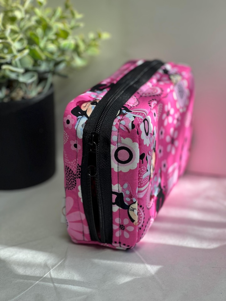 Op de afbeelding: Een rechthoekige roze make-up tas met een zwarte ritssluiting. De tas is versierd met een bloemenpatroon en stripfiguren. De tas is gemaakt van stof.