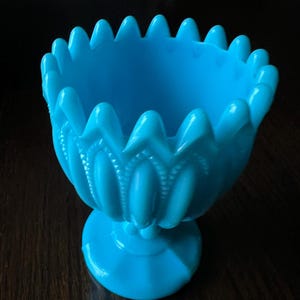 May include: A bright turquoise blue glass goblet featuring a scalloped, pointed rim and a vertical ribbed pattern along the bowl. The vessel stands on a circular pedestal base, showcasing a smooth, opaque finish with a consistent, vibrant color throughout.
