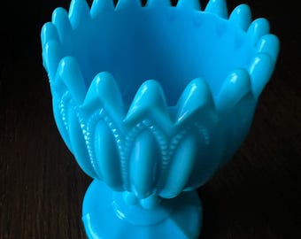 May include: A bright turquoise blue glass goblet featuring a scalloped, pointed rim and a vertical ribbed pattern along the bowl. The vessel stands on a circular pedestal base, showcasing a smooth, opaque finish with a consistent, vibrant color throughout.