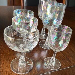 May include: A collection of six iridescent wine glasses and champagne flutes. The clear glass features a textured pattern and a shimmering, rainbow-like effect. The glasses are arranged on a dark wooden surface.