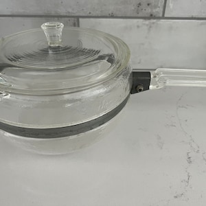 May include: A clear glass saucepan with a lid and a clear plastic handle. The saucepan has a silver band around the middle.