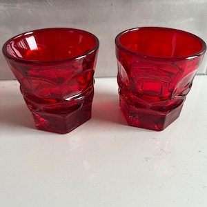 May include: Two red glass shot glasses with a textured design. The glasses are empty and sitting on a white surface.