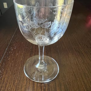 May include: A clear glass goblet with a wide bowl and a slender stem. The bowl is etched with a floral design, likely poppies, and sits on a round base. The glass is transparent, allowing light to pass through.