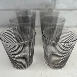 May include: Set of four clear drinking glasses with vertical black stripes. The glasses have a slightly angled design and are made of clear glass. The glasses are on a white surface.