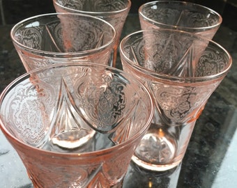 May include: Set of five pink glass tumblers with a delicate etched floral pattern. The glasses are clear with a light pink hue and have a slightly flared shape.