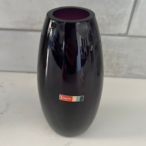 May include: A dark purple glass vase with a rounded bottom and a slightly flared top. The vase has a label that reads "Fostoria" on the front.