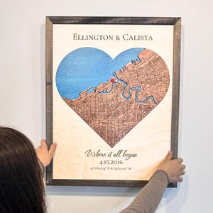 May include: A framed wooden map in the shape of a heart, featuring a detailed depiction of a city with blue water and brown land. The text "Ellington & Calista" is at the top, with "Where it all began" and a date below.