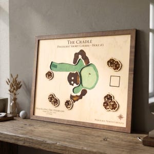 May include: Wooden framed artwork depicting a golf course hole. The map features green, brown, and tan tones. Text on the artwork reads "The Cradle", "Pinehurst Short Course - Hole #3", and "Hole-In-One Eric Heideloff".