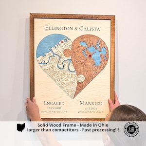 May include: Wooden framed artwork featuring a heart-shaped design with two puzzle piece sections. One section depicts a blue waterway, the other a brown landmass. Text includes "Ellington & Calista," "Engaged," and "Married" with dates and coordinates.