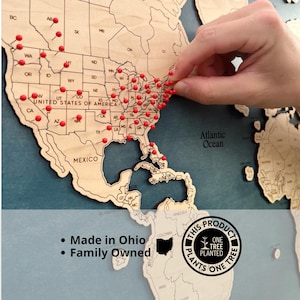 May include: A wooden world map with red push pins marking locations. The map is made of light-colored wood and is mounted on a blue background. The text "Made in Ohio" and "Family Owned" is visible. The product plants one tree.
