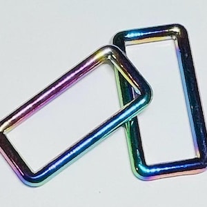 May include: Two iridescent rectangular metal rings. The rings have a rainbow finish, with colors shifting from purple to blue and green. The rings are positioned on a white surface.