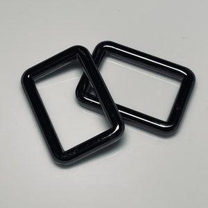 May include: Two glossy black rectangular metal rings. The rings are slightly angled, with rounded corners and a smooth, reflective surface. They are positioned on a white background, creating a contrast that highlights their shape and color.